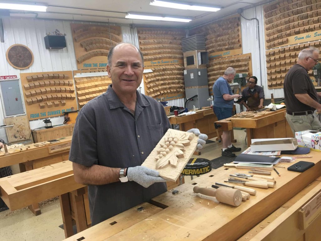 A person proudly holding a carved wooden piece in a workshop full of woodworking tools. The background shows various carved designs and enthusiastic participants engaging in wood carving classes, surrounded by workbenches and tools like chisels and mallets. This scene captures the essence of hands-on wood carving lessons, showcasing creativity and craftsmanship.