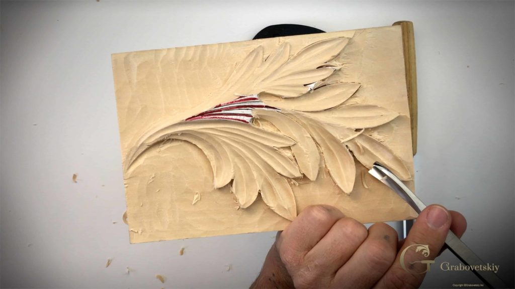 A hand holding a carving tool works on a detailed floral design in wood, showcasing the artistry of wood carving classes and lessons. The intricate leaves and lines come to life in the carved surface. Perfect for anyone looking to learn or improve their wood carving skills!!!