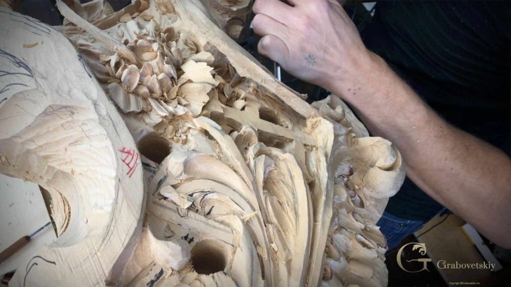 A close-up of a person's hand working on a detailed wood carving piece. You can see the intricate patterns taking shape with small tools, showcasing precision in the carving process. Shavings from the wood are scattered around, highlighting the craftsmanship involved in wood carving. This image could resonate with anyone interested in wood carving classes or lessons that focus on refining skills and techniques.