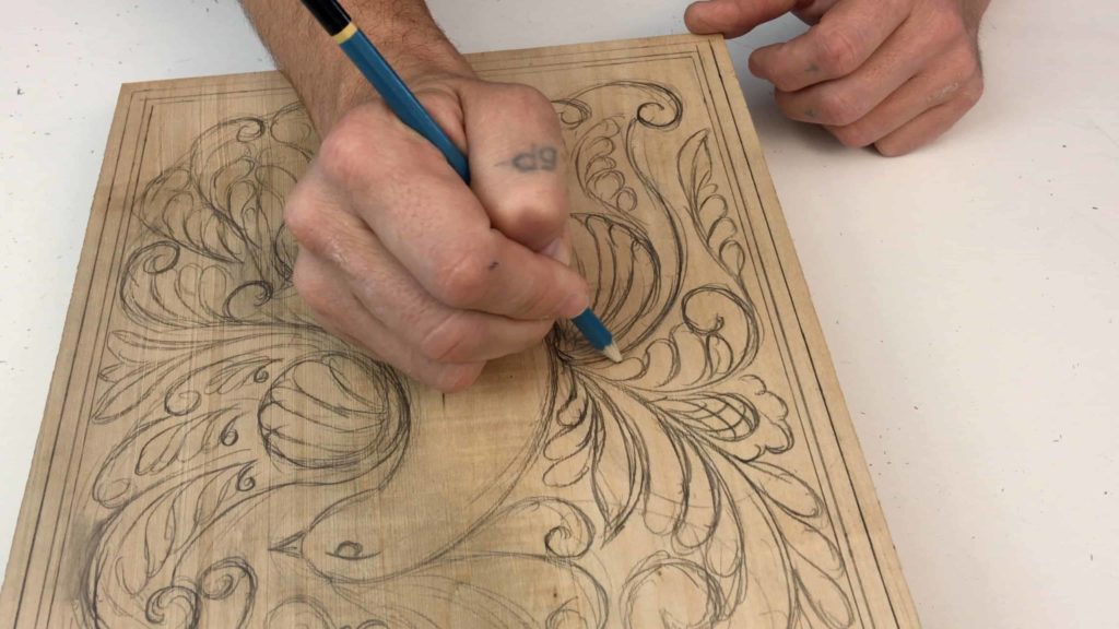 A person is sketching a detailed design on a piece of wood, preparing for wood carving classes. The outlines are intricate, showcasing the beginning of an artistic project. The hand is holding a pencil, likely planning the next steps in their wood carving lessons.