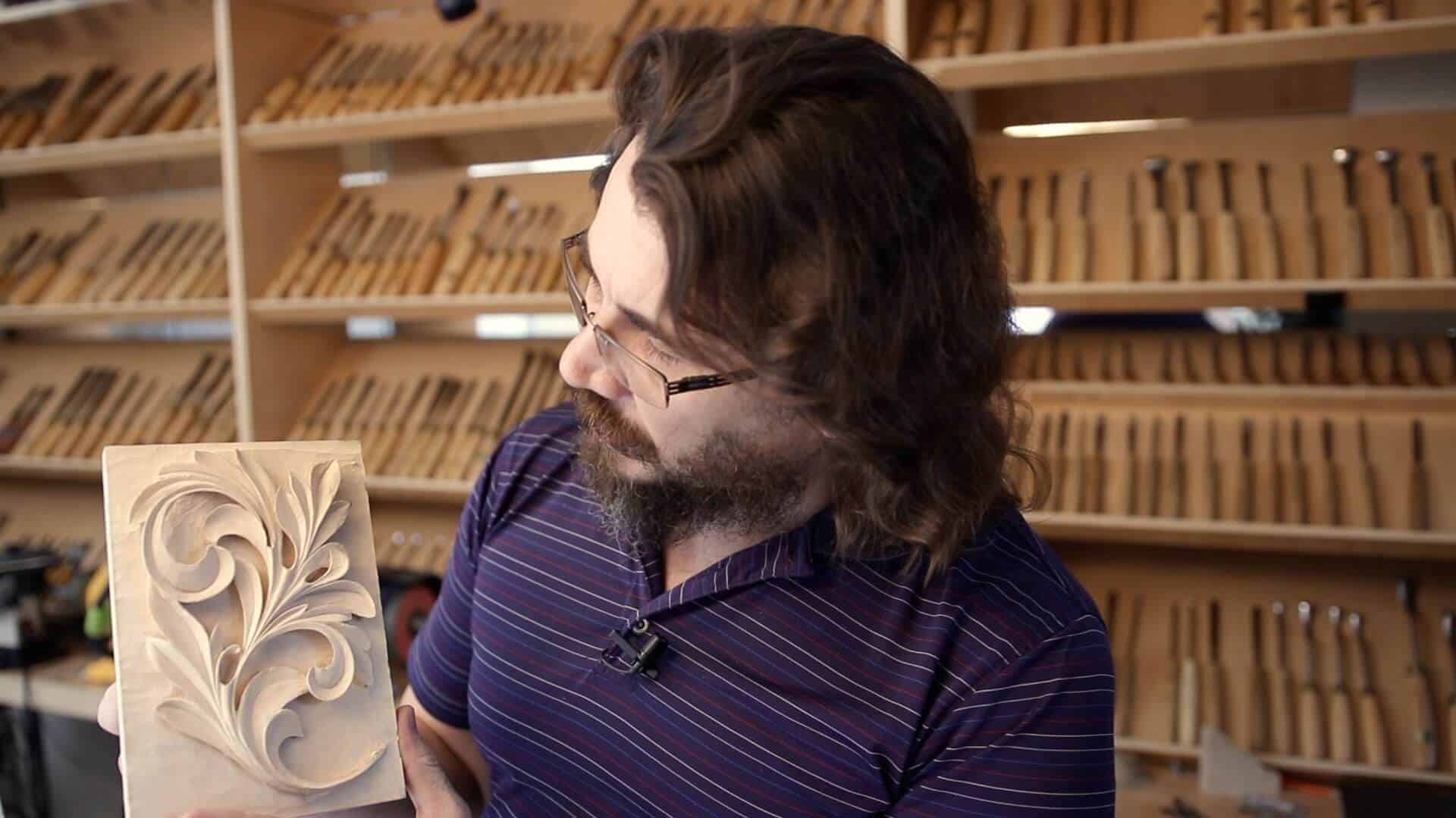 A guy holding a beautifully carved wood piece with intricate leaf patterns, surrounded by a wall of various wood carving tools. He seems really passionate about his work. Perfect for those looking for wood carving classes or lessons!