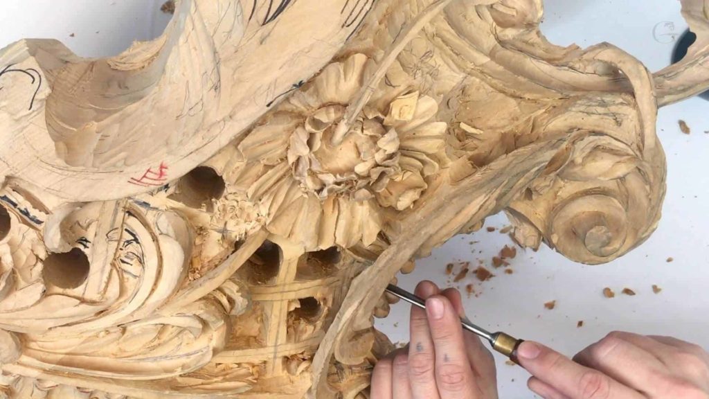 A close-up of a beautifully detailed wood carving in progress. You see a hand skillfully using a carving tool on intricate designs, including floral patterns and swirls. The wood has a light color, showing fine shavings scattered around, highlighting the artistic effort. This image perfectly captures the essence of wood carving classes and lessons, showcasing the dedication and craft involved in bringing wood to life.