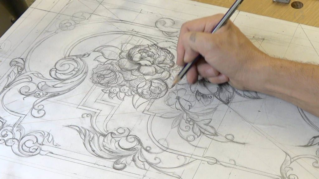 A person is sketching an intricate floral design on paper, preparing for wood carving. Their hand holds a pencil, and the artwork displays swirling lines, flowers, and decorative elements. This setting hints at the artistry involved in wood carving classes and lessons.