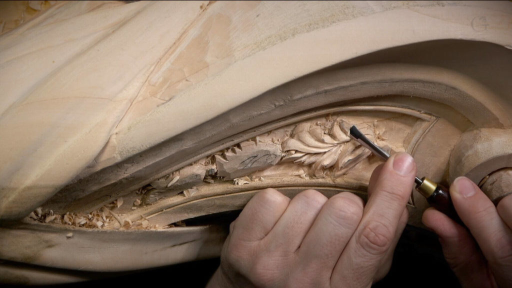WOODCARVING LESSONS: Woodcarver Grabovetskiy's hands are shown using a carving tool on a wooden sculpture. The close-up image captures the detailed work on the floral designs. - Wood Carving tutorials