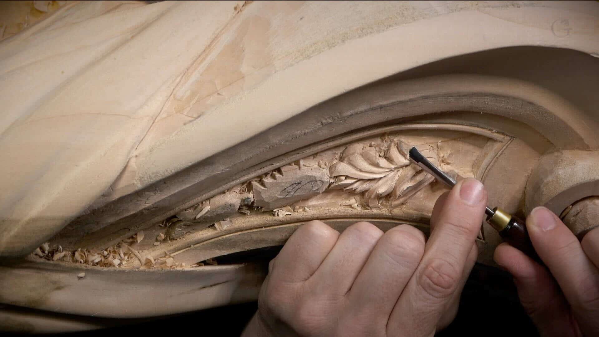 WOODCARVING LESSONS: Woodcarver Grabovetskiy's hands are shown using a carving tool on a wooden sculpture. The close-up image captures the detailed work on the floral designs. - Wood Carving tutorials