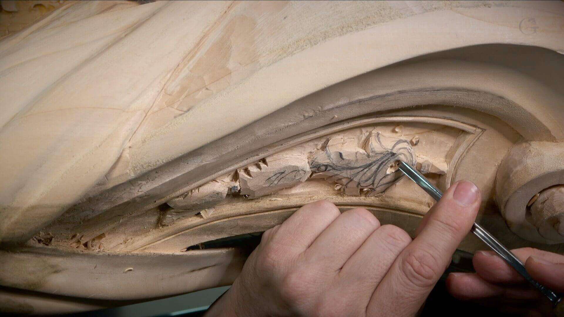 Intricate Wood Carving in Progress The image shows a close-up of a hand holding a carving tool, working on intricate wood carvings. The wood surface features detailed, flowing designs that appear to be in the process of being refined or completed. The focus is on the craftsmanship involved in wood carving.