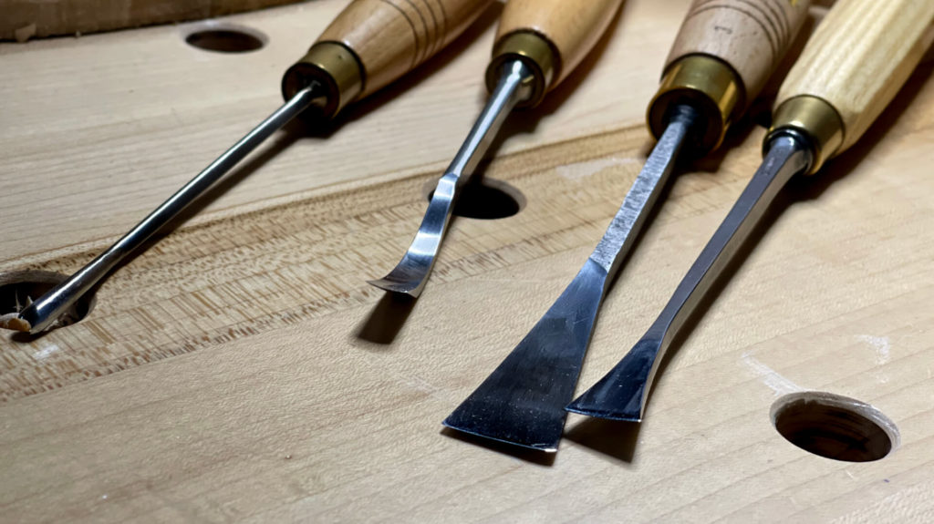 WOODCARVING LESSONS: Woodcarver Grabovetskiy shows four close-up woodworking tools with wood handles on a workbench with round holes. This kit includes a chisel and carving tools, perfect for beginners. Learn the best wood carving tools to start with today! - Wood Carving tutorials