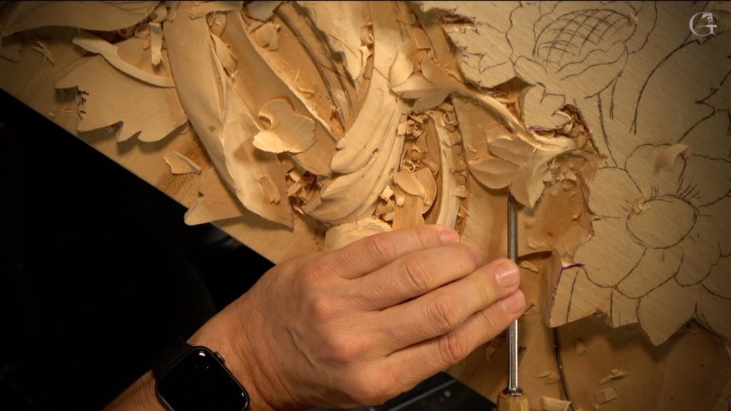 A close-up view of a person's hand skillfully carving details into a wooden surface. The carving tool is visible, creating intricate patterns and floral shapes. Shavings litter the workspace, highlighting the wood carving process. Perfect for showcasing wood carving classes and lessons!