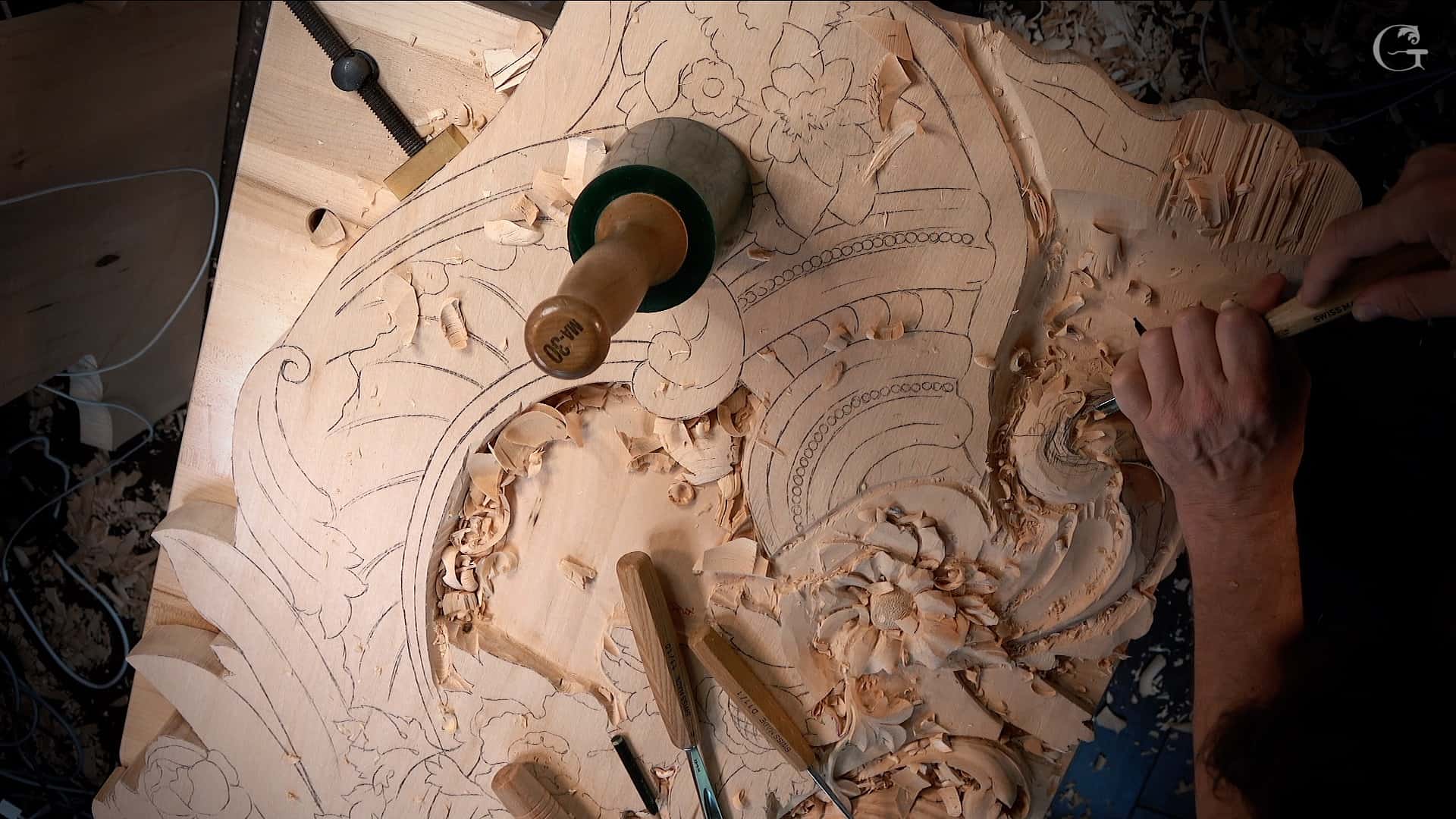 A close-up shows a wood carving in progress, featuring an intricate floral design. The scene displays a carving workbench with several tools, including a green-handled mallet and various chisels, surrounded by wood shavings. This setup highlights the creative process of wood carving, a skill emphasized in wood carving classes and lessons.