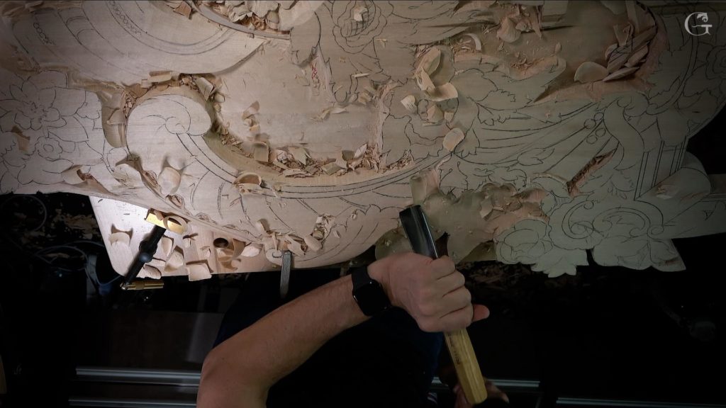 A close-up shot of a wood carving in progress shows intricate designs taking shape. The craftsman hammers a chisel into the wood surface, surrounded by shavings and curls from the work. This image captures the essence of wood carving lessons, showcasing skill and artistry that come with dedicated practice in wood carving classes.