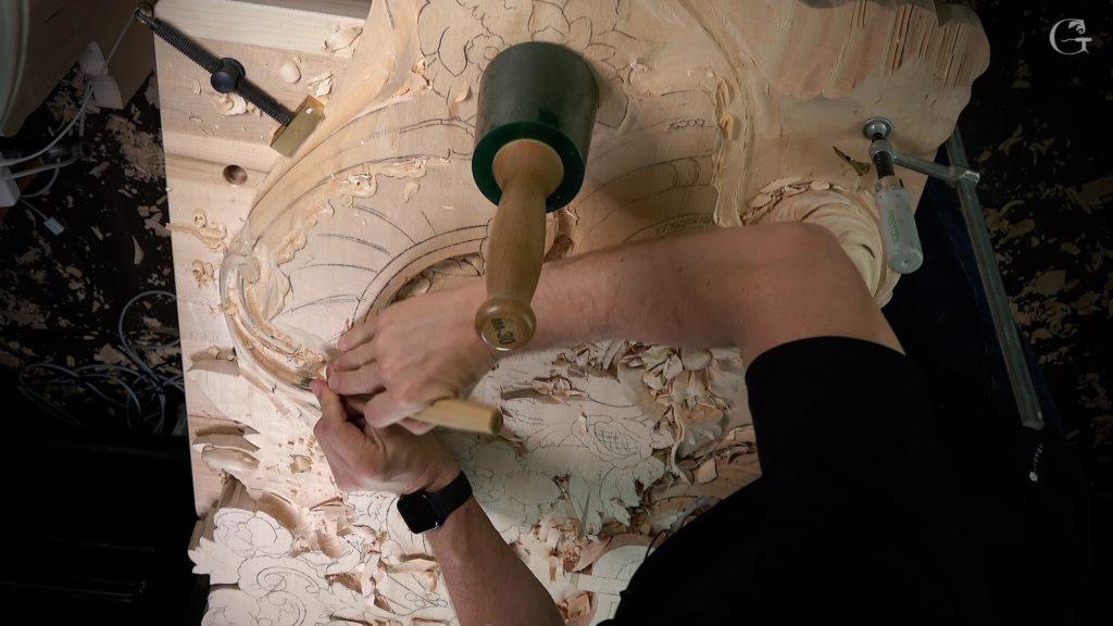 A person carving a detailed wooden design on a workbench. They're using a mallet and a chisel to shape the wood. Shavings cover the surface, showcasing the beauty of the carving process. This scene captures the essence of wood carving classes and lessons, highlighting skill and creativity in action.