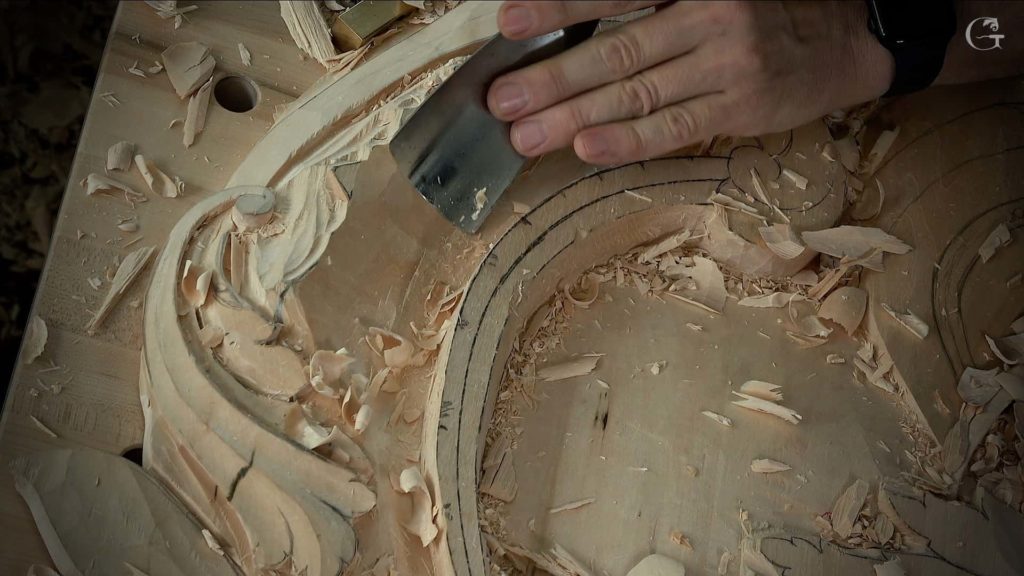 A close-up of a hand using a carving tool on a wooden surface. The wood shows fine shavings and a detailed design in progress. This scene highlights the art of wood carving, perfect for showcasing in wood carving classes or lessons.