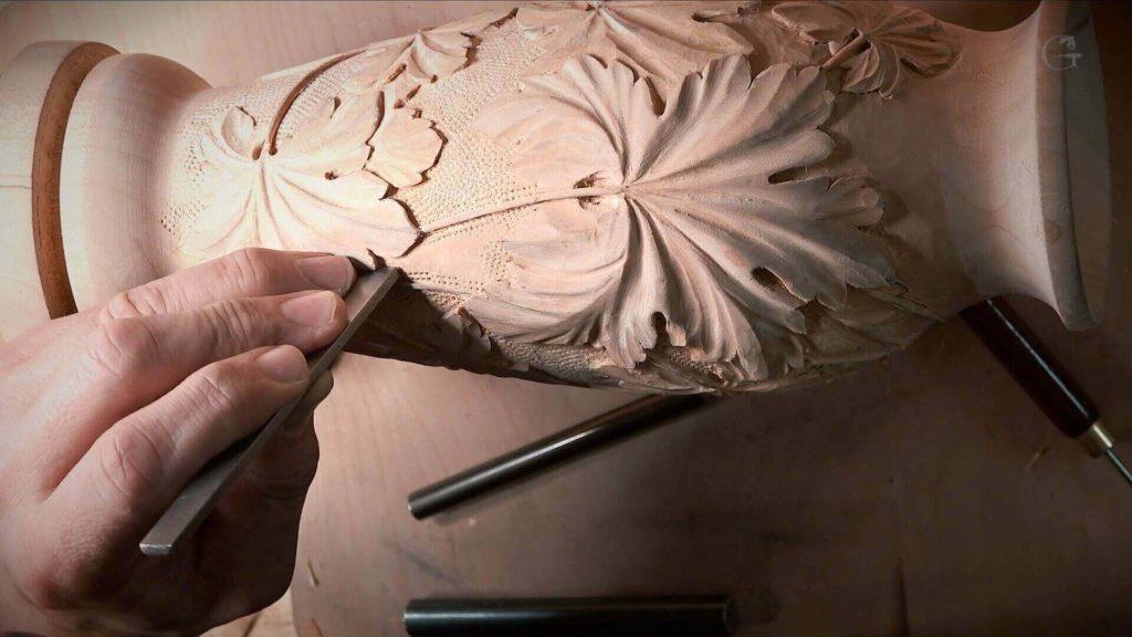 A close-up view of a hand guiding a chisel along a beautifully carved wood vase, showcasing intricate leaf designs. The scene highlights a dedicated woodworker at work, using chisels and tools on a sturdy workbench, embodying the passion behind wood carving. Perfect for those interested in wood carving classes and lessons!