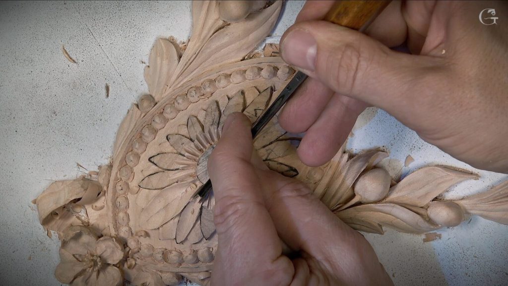 Alt text: A close-up of hands carving a detailed floral design in wood with a chisel. This image represents the art of wood carving and highlights the precision involved in wood carving classes and lessons.