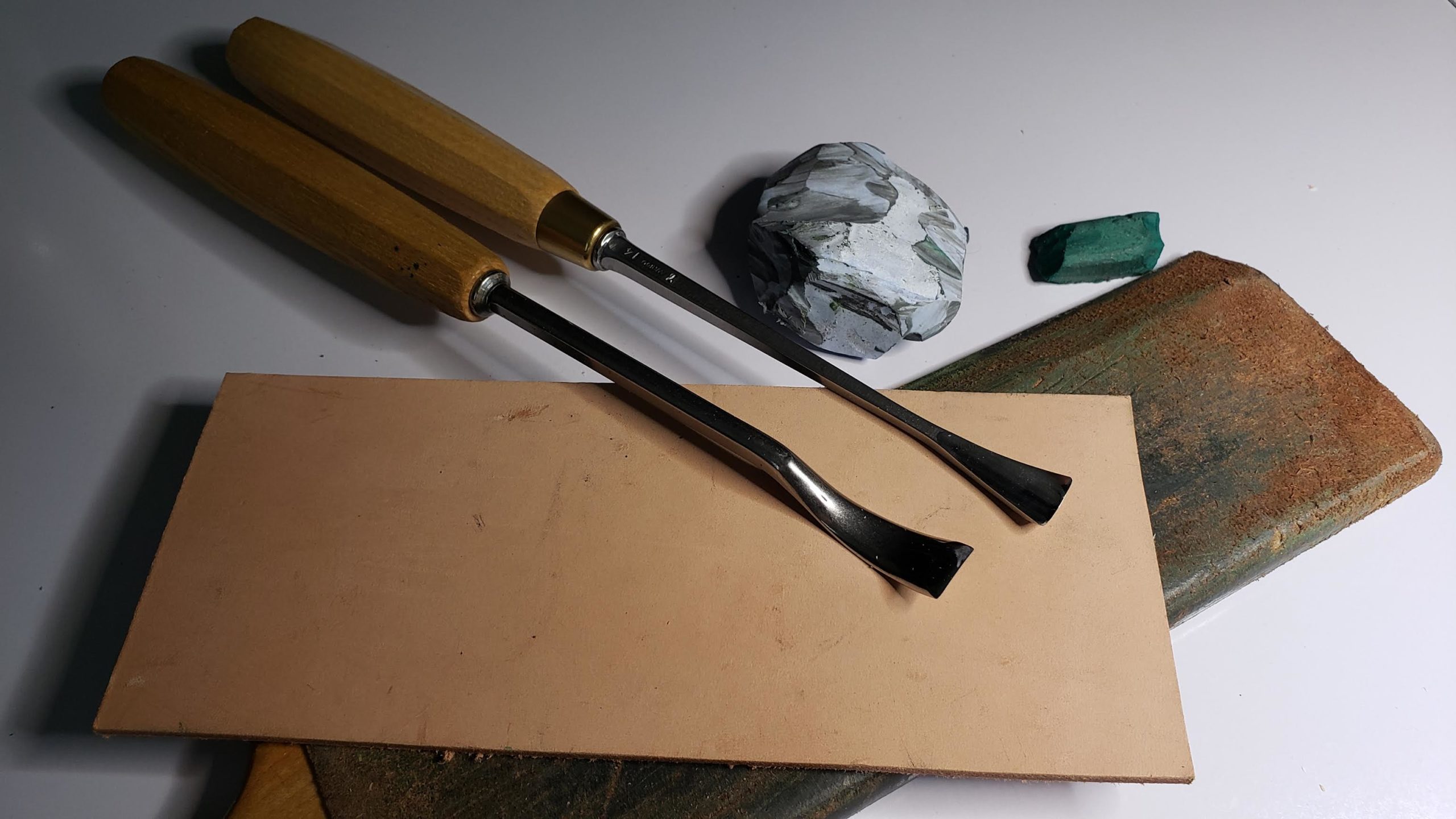 Two wood carving tools rest on a light-colored work surface, alongside some modeling clay and a wooden carving board. The tools feature wooden handles and shiny metal blades, ready for action. Perfect for anyone interested in wood carving classes or lessons!