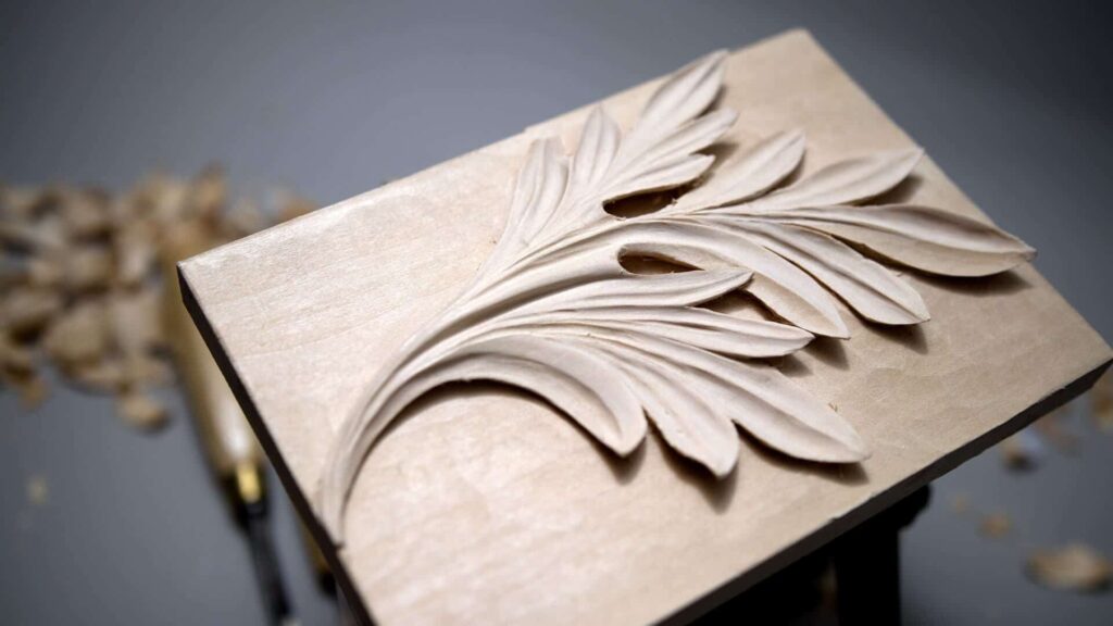 Alt text: A beautifully carved design of a leaf pattern on a piece of light-colored wood, showcasing the details of the carving. Shavings and tools are scattered in the background, highlighting the wood carving process and the art of wood carving classes and lessons.