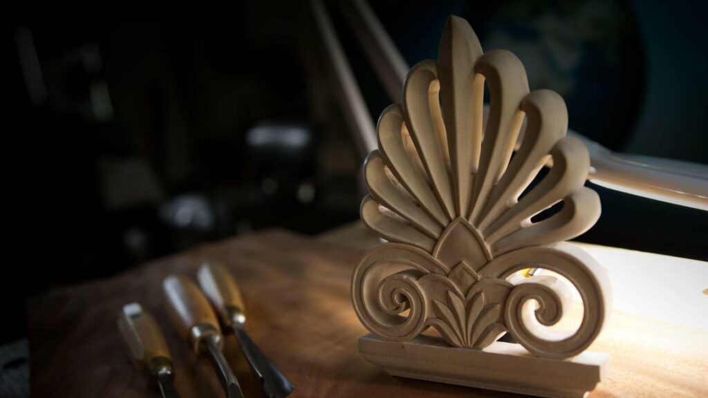 Alt text: "A beautifully carved design made from light wood, showcasing elegant curves and details, sits on a workbench beside sharp wood carving tools, emphasizing the beauty of craftsmanship. Perfect for those interested in wood carving classes and lessons."