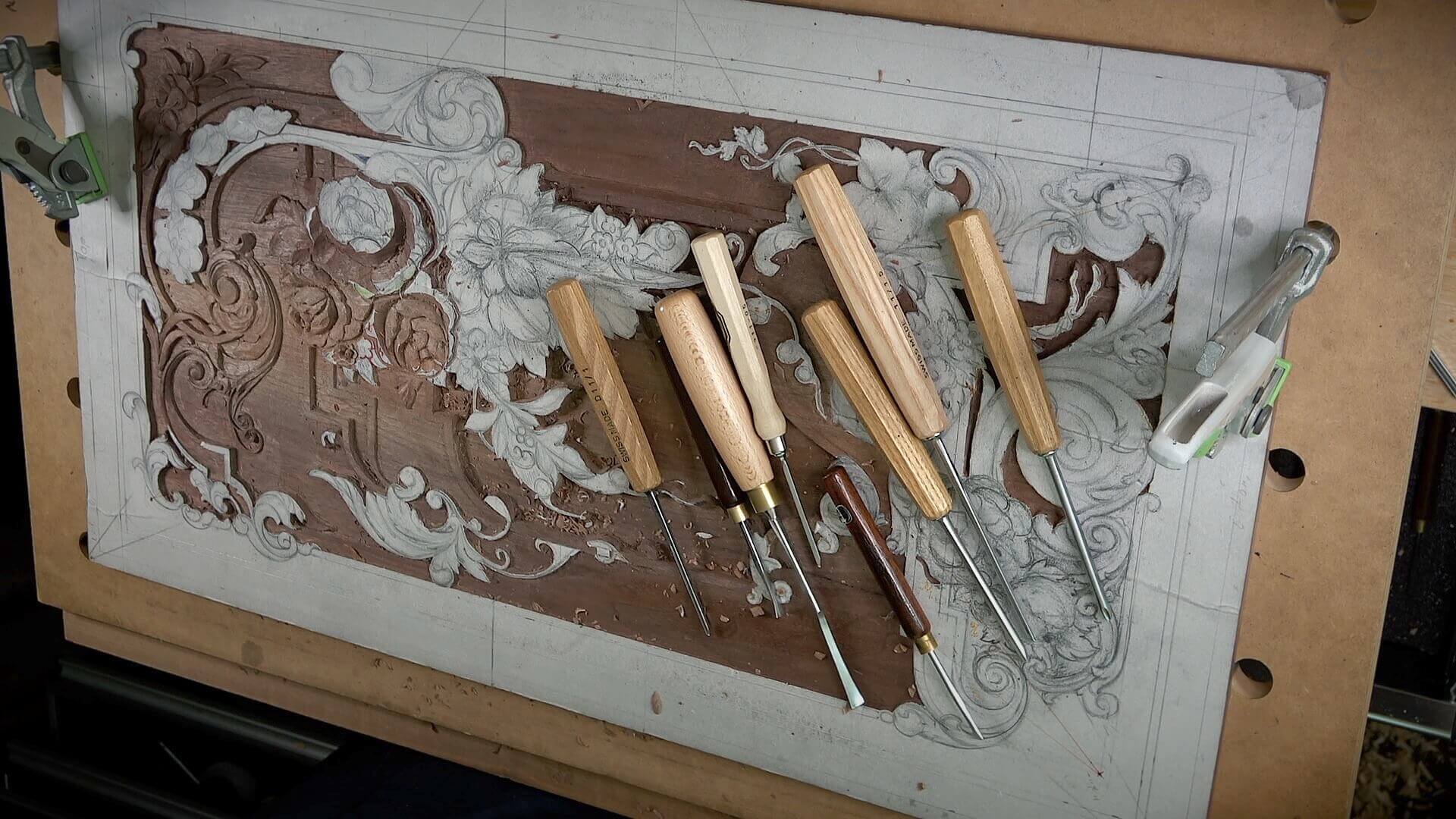 A workbench filled with various wood carving tools is shown, alongside a beautifully detailed wood panel featuring an intricate floral design. This setup highlights the art of wood carving, perfect for those interested in wood carving classes or lessons. The tools, including chisels and gouges, suggest dedication to this craft, encouraging aspiring artists to harness their creative skills. Happy carving!!!
