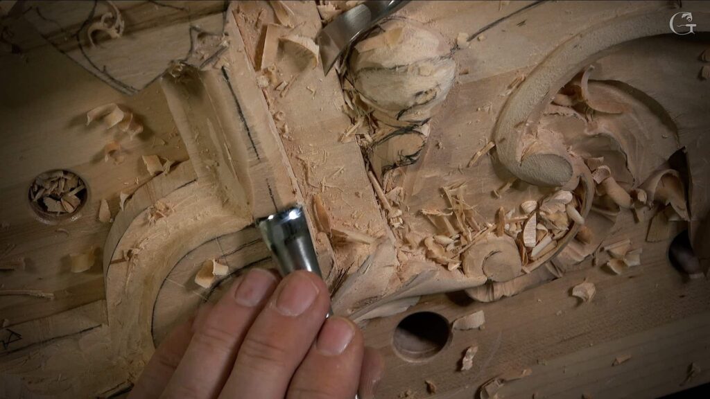A hand is holding a carving tool, working on a piece of wood. There are shavings scattered around, showing recent carving. You can see some detailed patterns emerging in the wood, hinting at a project in progress. This scene perfectly captures the crafting vibe, ideal for wood carving classes or lessons for anyone interested in honing their skills!