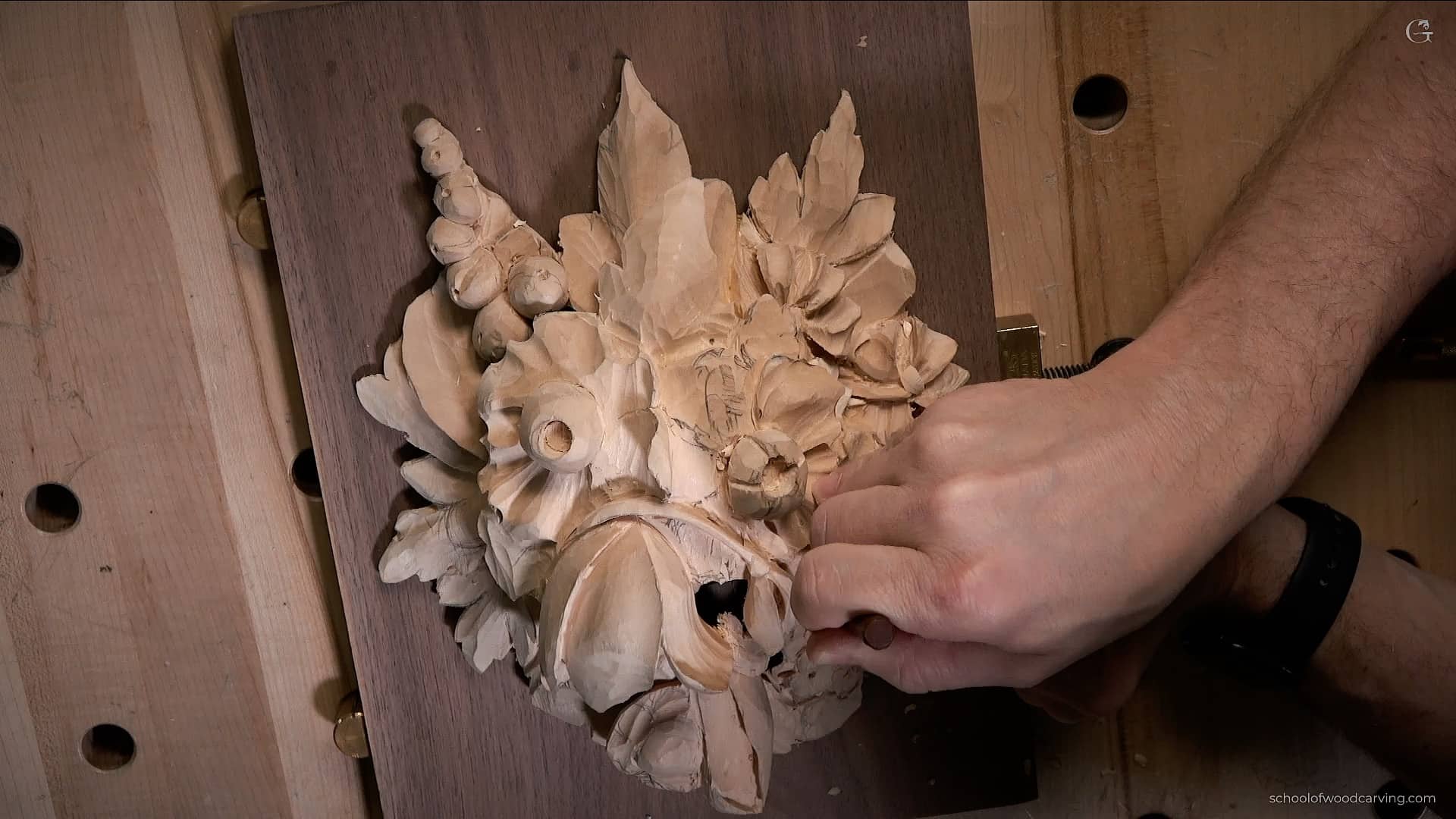 A person is carving a detailed floral design in wood on a workbench, showing their hands and tools in the process. This image captures a moment from wood carving classes or lessons, highlighting the skill and creativity involved in the craft.