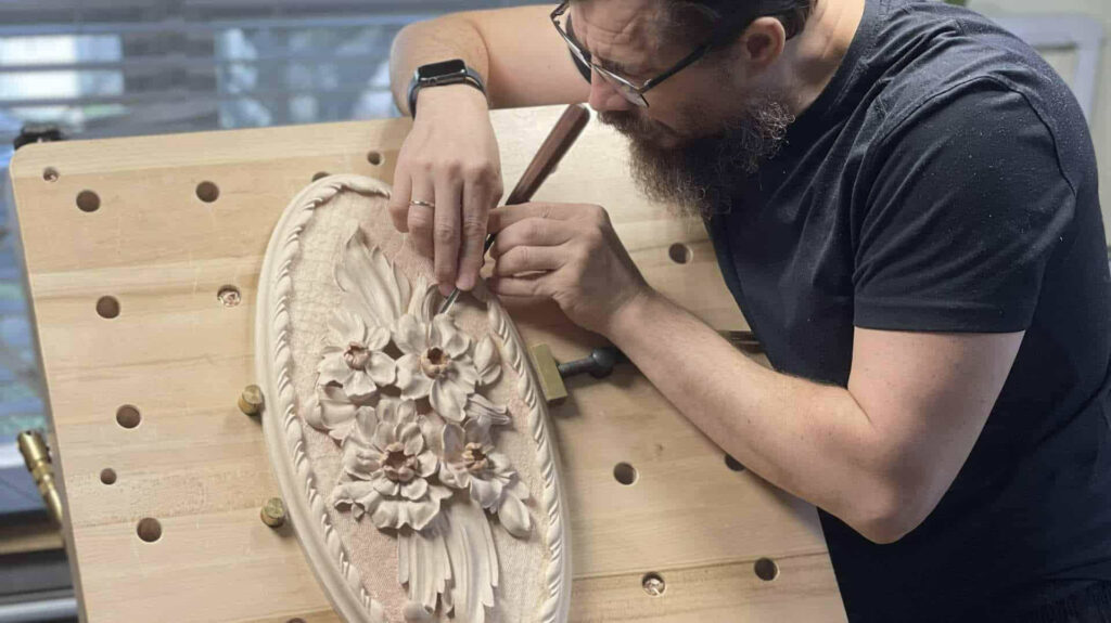 A wood carving artist focuses intently on a detailed floral design on a carving workbench, using a gouge tool. The craftsmanship captures the beauty of each flower, showcasing skills learned in wood carving classes and lessons. Dust motes float in the light, hinting at hours spent honing these techniques.