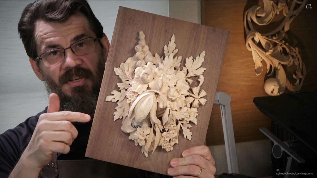 A bearded man wearing glasses is pointing at a detailed floral wood carving mounted on a rectangular wooden board that he is holding up.
