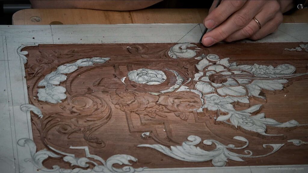 A person is focused on carving a detailed floral design on a piece of wood. They're using a carving tool, delicately working on the intricate patterns. The wood shows both darker and lighter areas, highlighting the depth of the carving. This image captures the essence of hands-on wood carving lessons, perfect for anyone interested in wood carving classes!