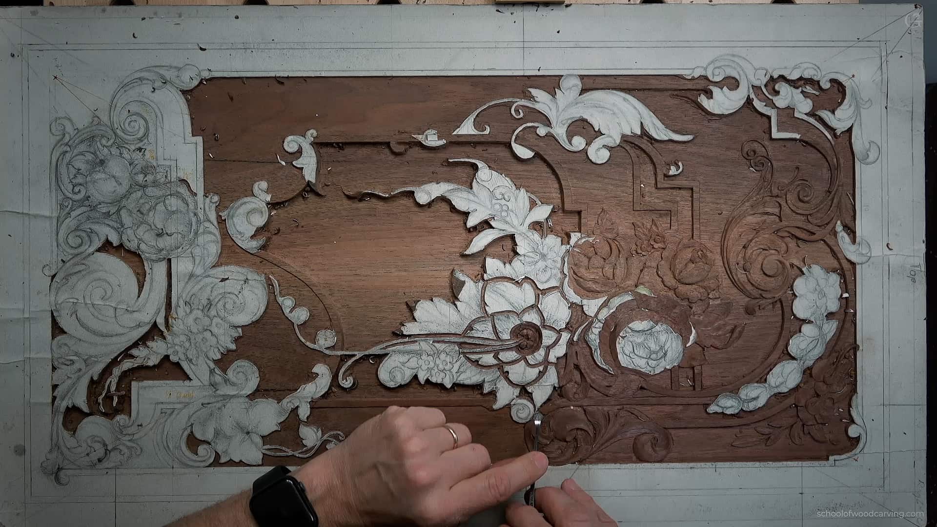 Hand carving detailed floral and scroll patterns on a wooden panel using a small carving tool.