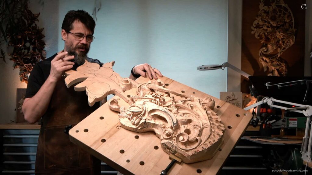 A wood carving instructor demonstrating a beautifully detailed floral design on a large wooden piece. The artist holds the carving with both hands, showcasing the intricate curves and shapes while surrounded by tools and materials in a well-lit workshop. Perfect for those looking for wood carving classes or lessons.