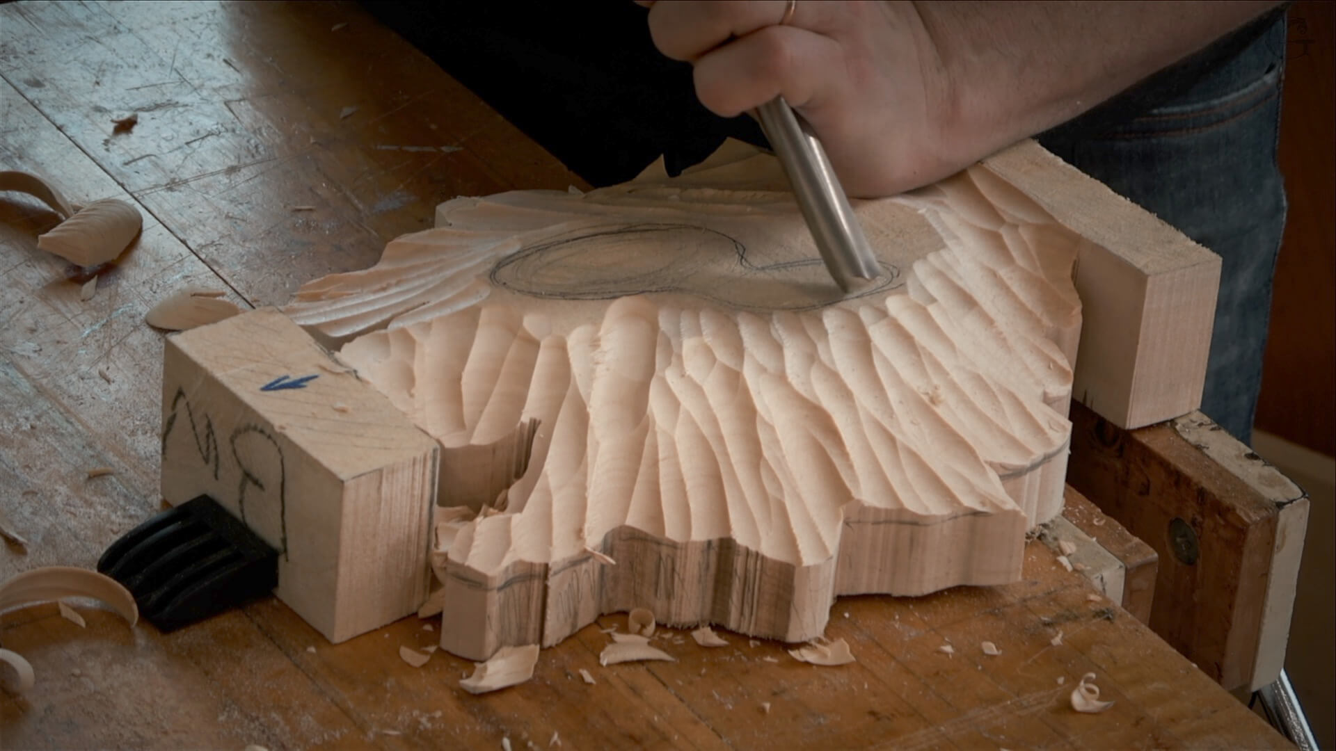 Close-up of a person using a chisel to carve a design into a piece of wood clamped to a workbench. Wood shavings are scattered on the surface around the carving.