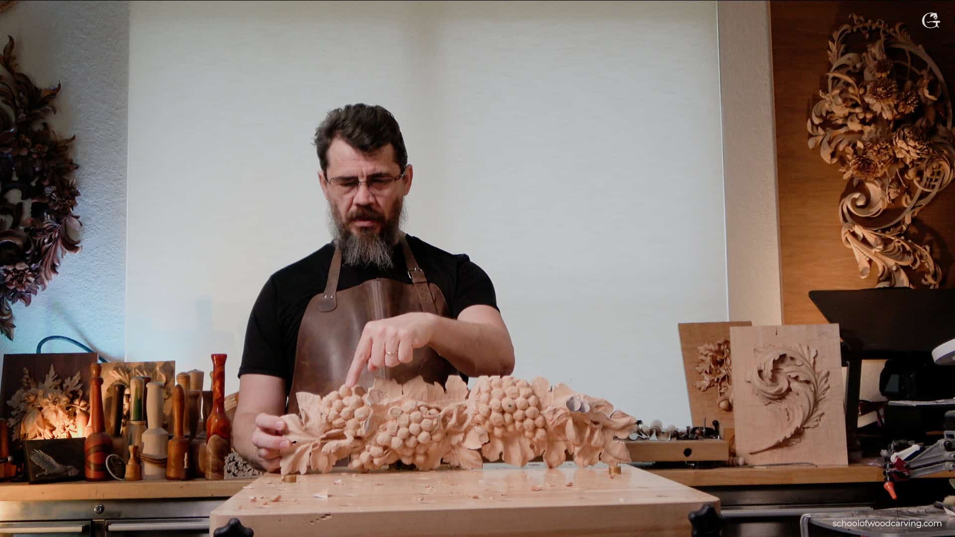 A person in a black t-shirt and brown apron is working on a detailed woodcarving of grapes on a table, surrounded by various woodworking tools and finished carvings, possibly inspired by woodcarving tutorials online.