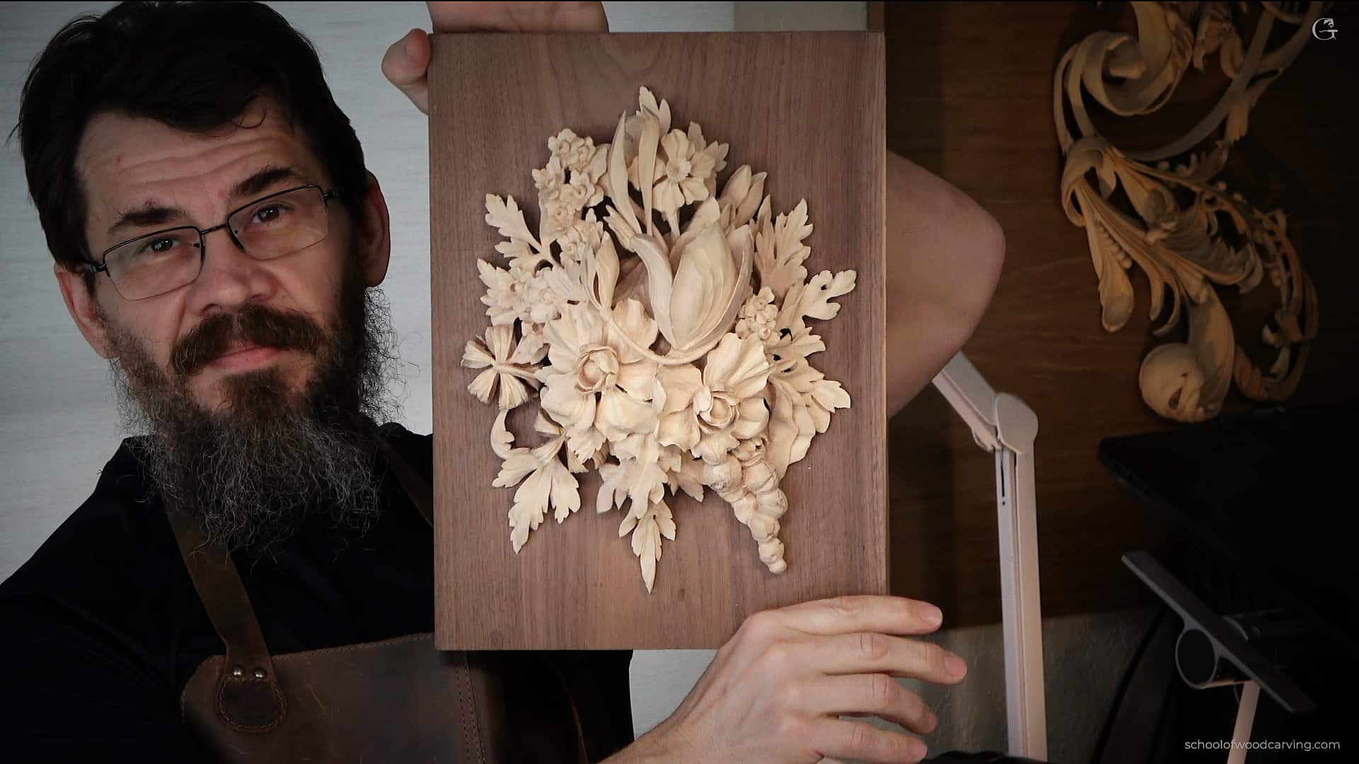 A man with glasses and a beard holds up a wood carving of flowers and leaves. He is wearing a leather apron, and the background includes more wooden carvings, reminiscent of the detailed projects you can learn in woodcarving tutorials online.