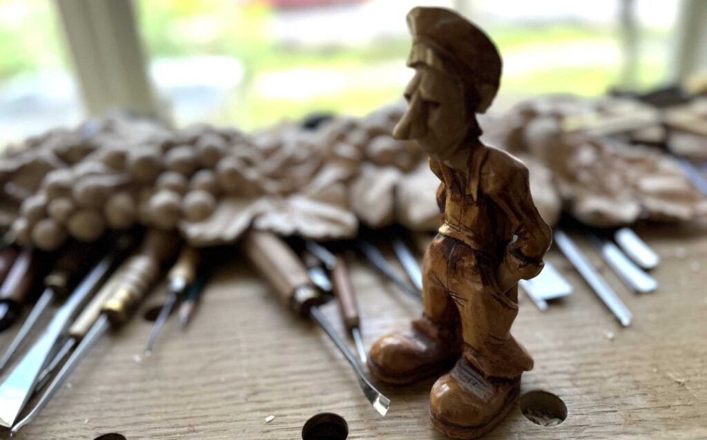 WOODCARVING LESSONS: A wooden carving of a man with a hat is on a workbench. Carving tools and wood shavings are around it. Woodcarver Grabovetskiy uses these tools. - Wood Carving tutorials