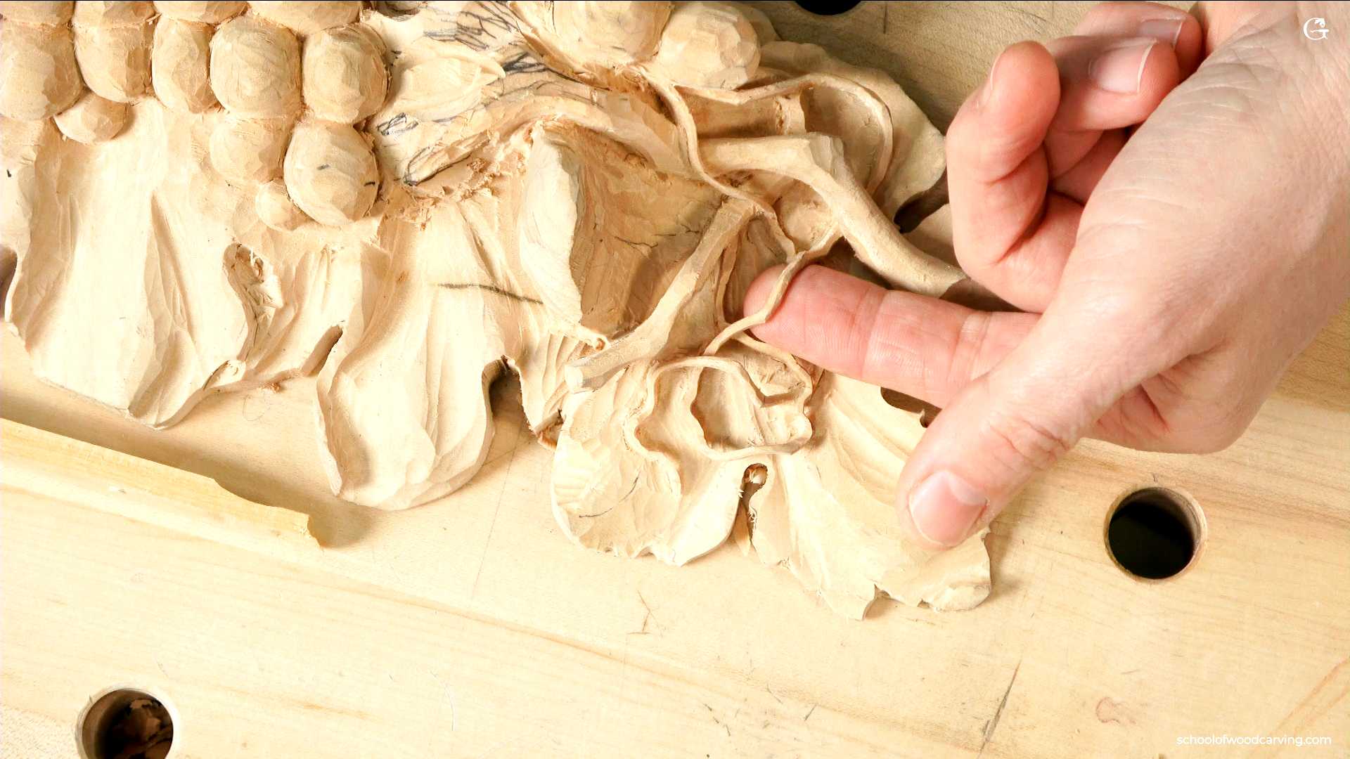 WOODCARVING LESSONS: Woodcarver Grabovetskiy uses his fingers to hold a detailed wood carving of leaves and grapes. The work is happening on a workbench. - Wood Carving tutorials