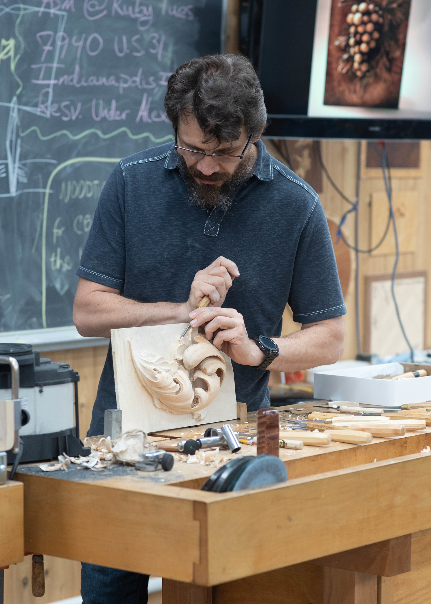 WOODCARVING LESSONS: Woodcarver Grabovetskiy, a bearded man, carves wood at his woodworking station. Tools are all around him. In the background, you can see a chalkboard and shop equipment. - Wood Carving tutorials