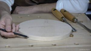 Hand holding a pen above a circular piece of wood on a carving workbench, surrounded by carving tools. Looks like a great setup for wood carving classes or lessons!