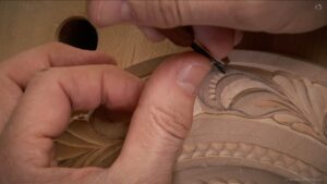 A person is carving a detailed design in wood, focusing intently while using a small carving tool. The setup shows a close-up of hands at work, with delicate lines starting to form in the wood surface. This image highlights the beauty and skill involved in wood carving, showcasing the kind of precision taught in wood carving classes and lessons.
