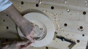 A person carving a design into a round piece of wood on a workbench. They're using a sharp carving tool, surrounded by wood shavings. This image highlights the hands-on nature of wood carving, perfect for showcasing wood carving classes and lessons.
