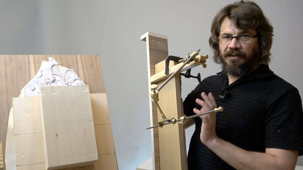 WOODCARVING LESSONS: Woodcarver Grabovetskiy, a man with glasses and a beard, shows how to use a pointing machine for exact measurements in his workshop. On the left side, you can see part of a wooden structure. - Wood Carving tutorials