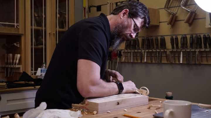 WOODCARVING LESSONS: Woodcarver Grabovetskiy, a man with a beard and glasses, works carefully on wood in his workshop. He uses carving tools on a wooden piece. Behind him, many woodworking tools are neatly arranged on the wall. - Wood Carving tutorials