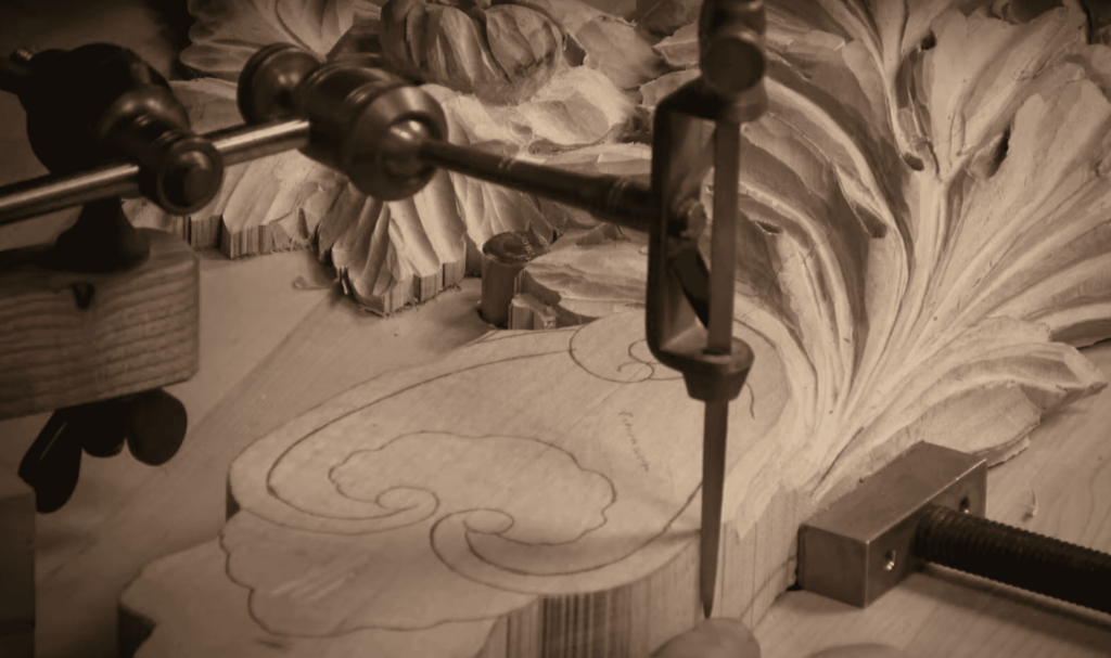 WOODCARVING LESSONS: A close-up of woodcarving shows detailed designs. A metal clamp holds the wood still while a tool carves. Woodcarver Grabovetskiy is in the picture. Learn how to use a pointing machine to copy these details exactly in your woodcarving projects. - Wood Carving tutorials