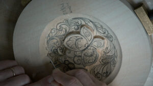 Alt text: A close-up of a hand carving a detailed floral design into a light-colored piece of wood. The design features flowing lines and patterns, showcasing the precision involved in wood carving. This image highlights the artistry in wood carving lessons, perfect for anyone interested in wood carving classes.