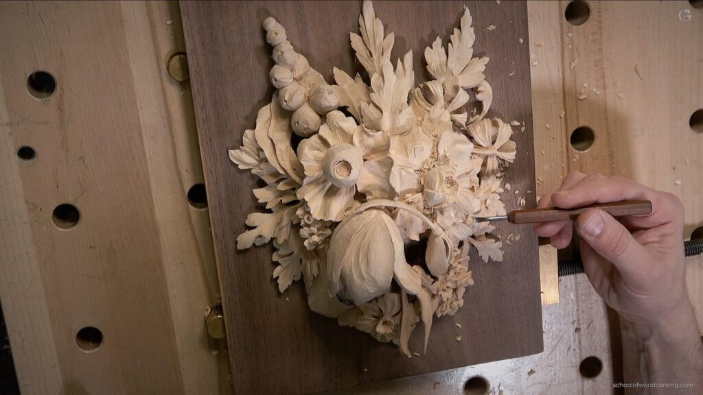 Alt text: A close-up of a wood carving project showing detailed floral and leaf designs being carved with a fine tool. The workbench is visible, highlighting shavings and a rich wood grain. Perfect for illustrating wood carving classes and lessons! Wood Carving Tutorials- Wood Carving Classes - Wood Carving Lessons - Wood Carving Video Lessons