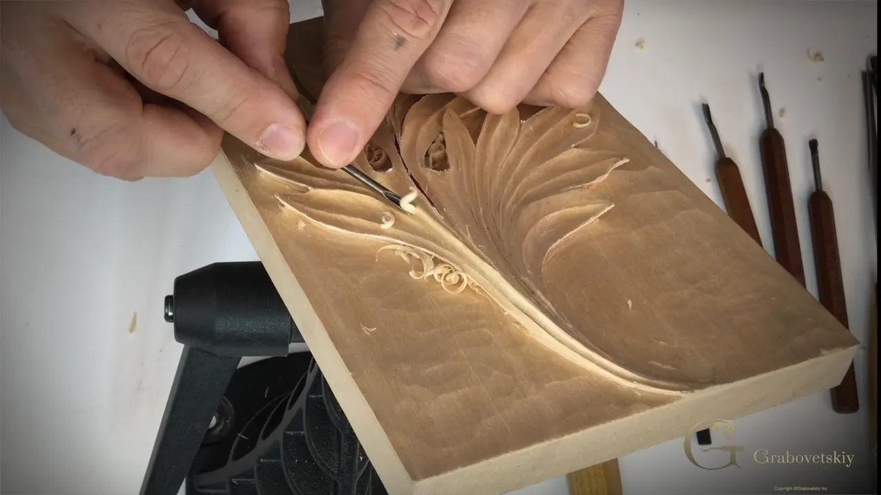 Both hands steer a small gouge along the finished lobes of the leaf, the panel standing on the bench.