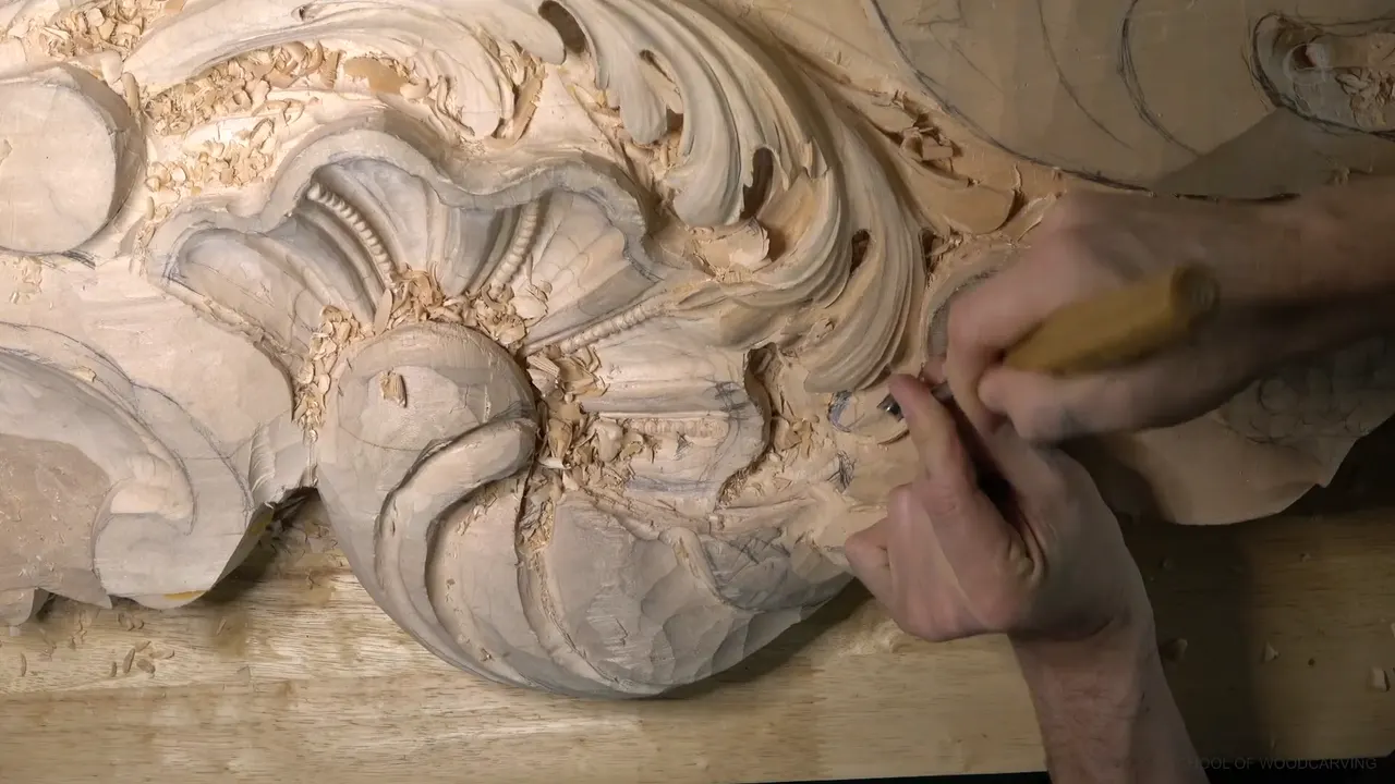 A yellow handled gouge pressed into an acanthus lobe beside the carved shell, both hands on the tool.