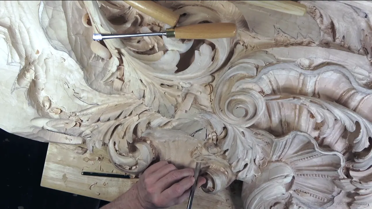 Acanthus leaves curling around a carved shell, seen from above, with a hand holding a gouge near the lower edge.