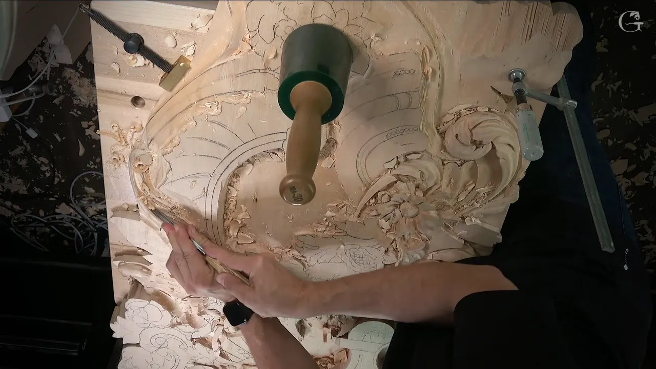 Both hands steering a small tool along the edge of a carved scroll, green mallet resting above