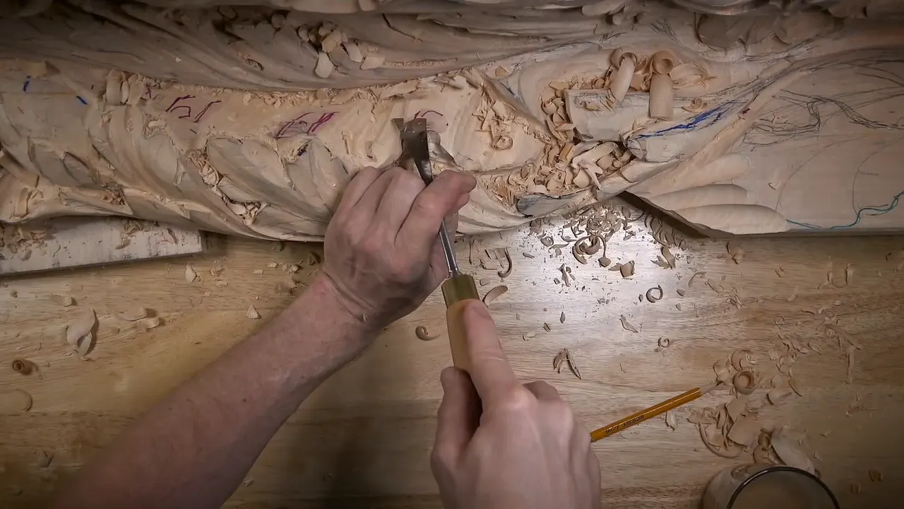 Overhead view of both hands driving a gouge into the lower edge of a carved relief, pencil on the bench