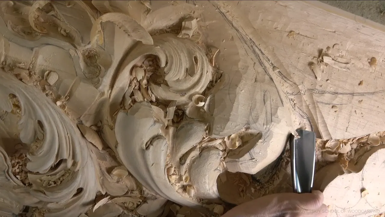 A wide gouge pushes into the curve beside a finished scroll, pencil lines still on the flat wood.