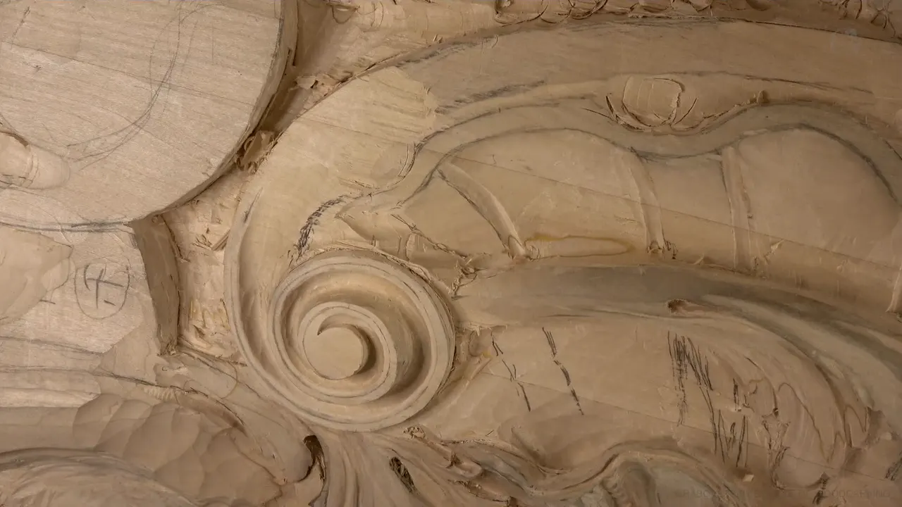 A carved volute in warm light, its spiral clean, drawn marks on the wood to its right.
